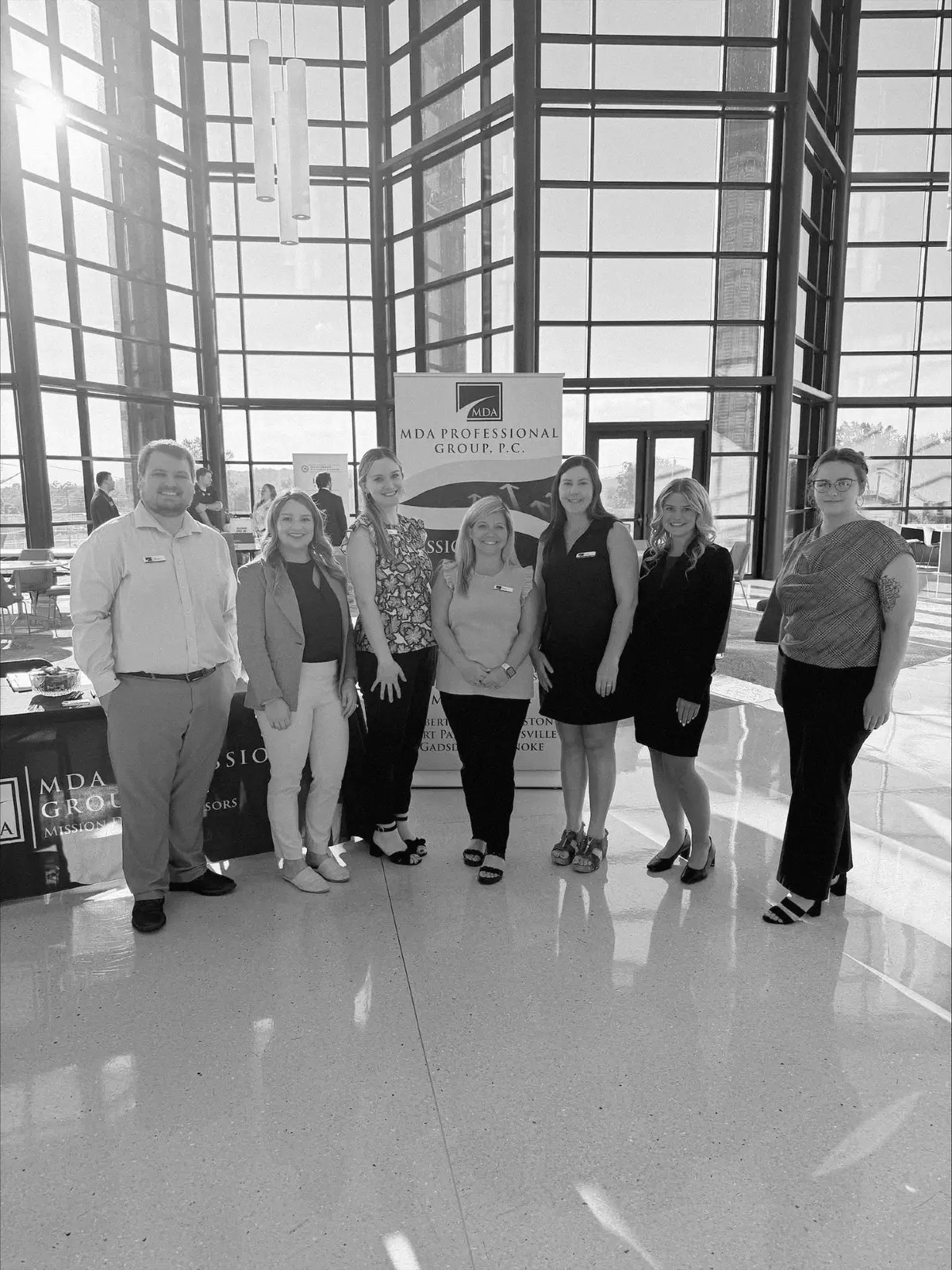 Seven MDA team members in front of the firm banner at a hiring event in a glass atrium